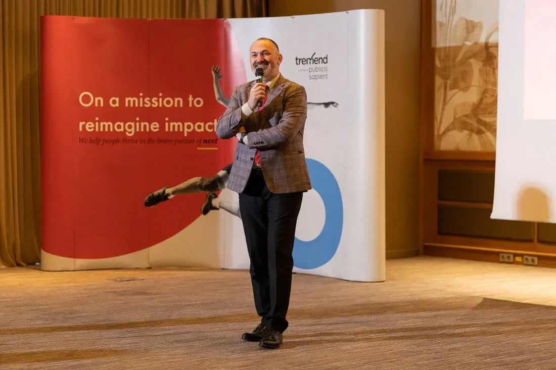 Un bărbat în sacou în carouri și cravată roșie vorbește la microfon în fața unui banner Tremend Publicis Sapient cu textul «On a mission to reimagine impact».