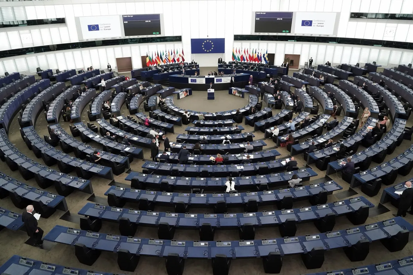 Vedere largă a hemiciclului Parlamentului European de la Strasbourg în timpul plenarei inaugurale a Conferinței privind viitorul Europei, cu rânduri de pupitre albastre care se curbează spre o tribună flancată de steaguri.
