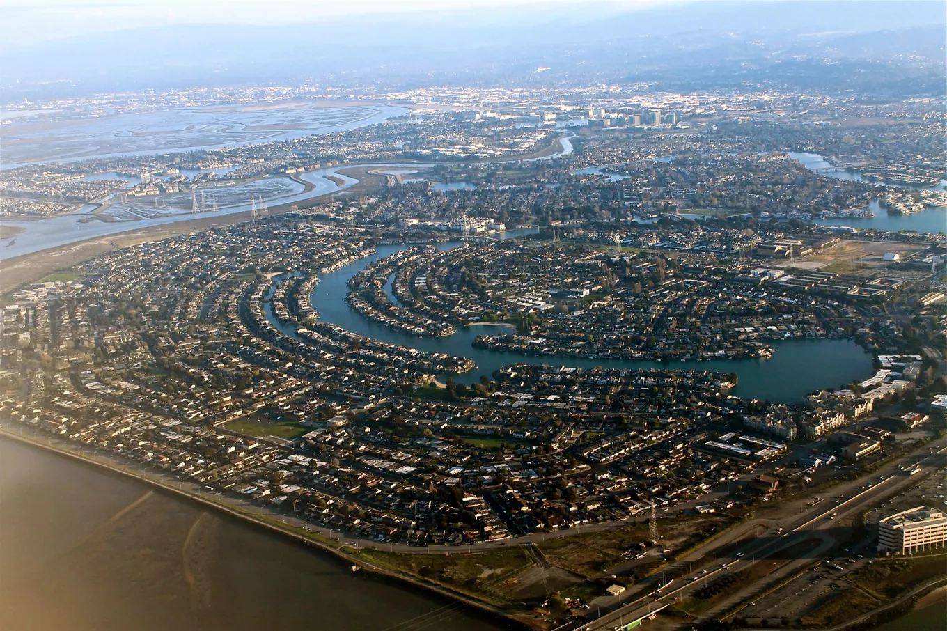Vedere aeriană asupra Silicon Valley: cartiere dense care se curbează în jurul unor lagune la marginea golfului San Francisco, cu valea întinzându-se până la dealuri.