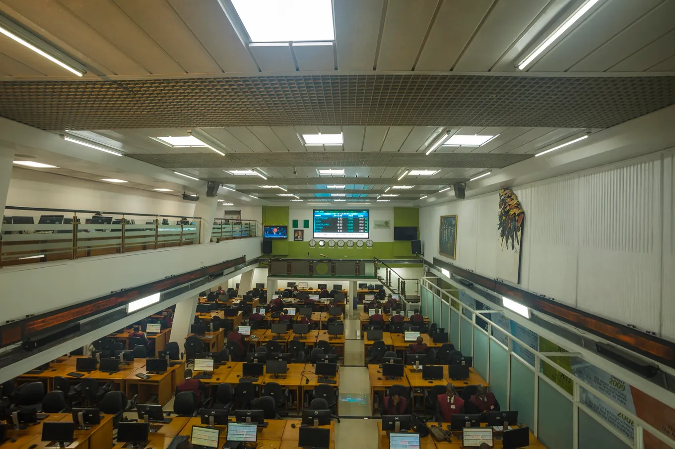 Sala de tranzacționare a unei burse văzută de la balcon: rânduri de birouri cu monitoare, brokeri la lucru și un panou cu cotații pe peretele din fund.