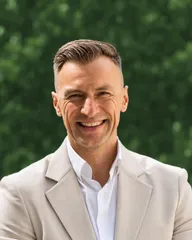 Marius Hanganu zâmbind, în sacou bej deschis și cămașă albă, cu frunziș verde în fundal.
