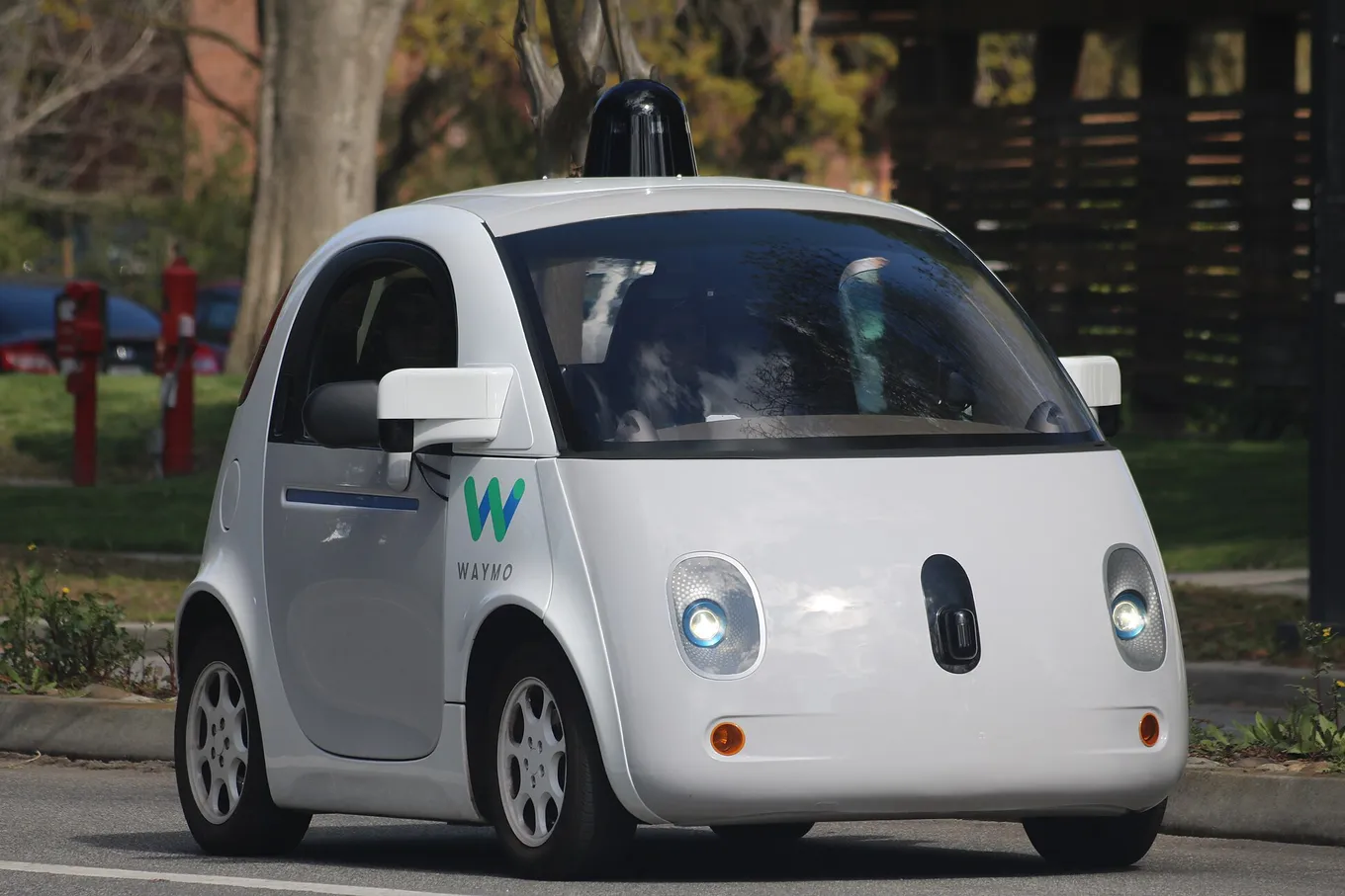 O mașină autonomă Waymo, mică și albă, cu un senzor în formă de dom pe acoperiș, pe o stradă mărginită de copaci.