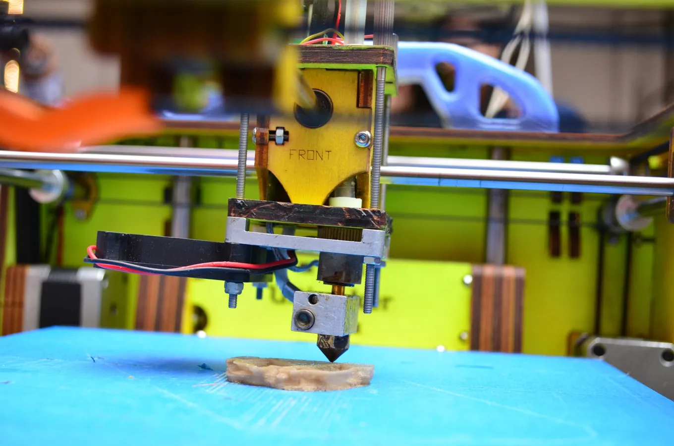 O imprimantă 3D în funcțiune: capul de printare depune primele straturi ale unei piese pe o placă albastră.