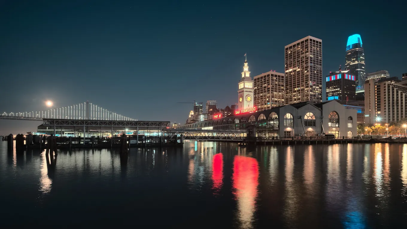 Clădirea Ferry Building și turnurile din centrul San Francisco noaptea, cu podul Bay Bridge luminat în stânga, oglindite în golf.