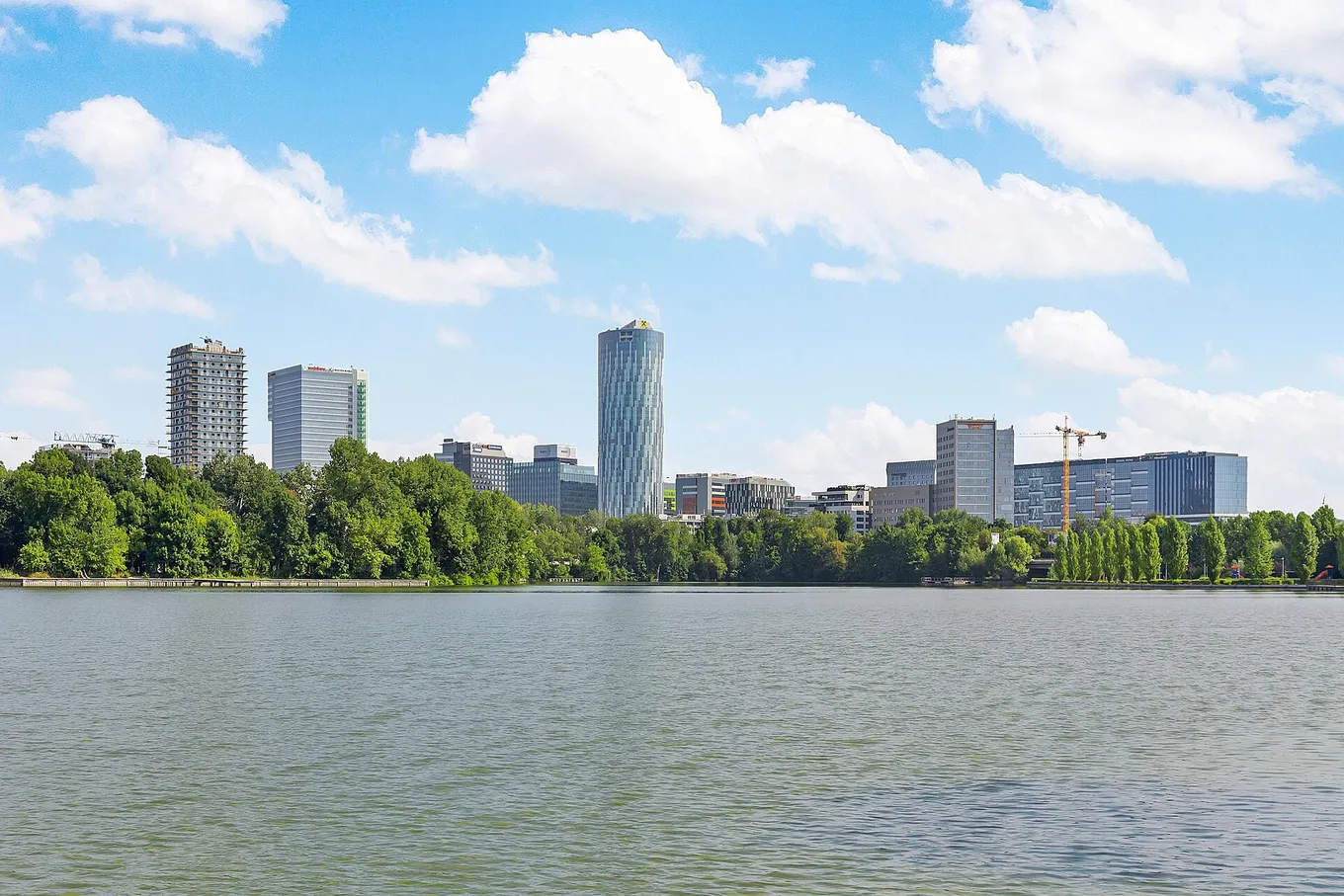 Clădirile de birouri din cartierul de afaceri Floreasca, București, văzute peste lacul Herăstrău, sub un cer albastru cu nori albi.