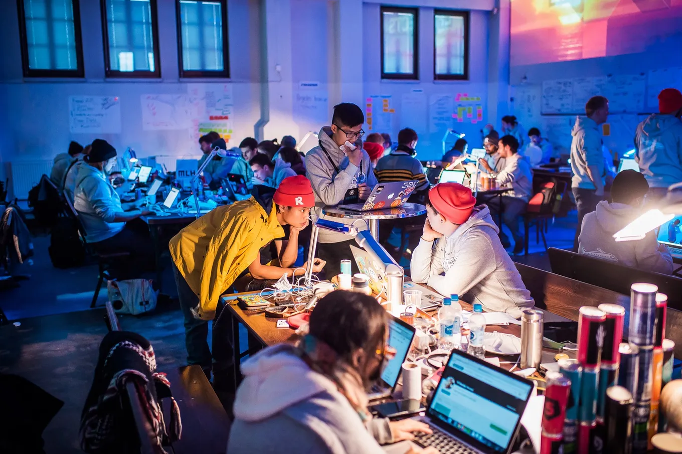 Programatori care lucrează noaptea pe laptopuri la mese lungi, într-o sală de hackathon aglomerată, luminată în albastru și violet, cu notițe prinse pe peretele din spate.