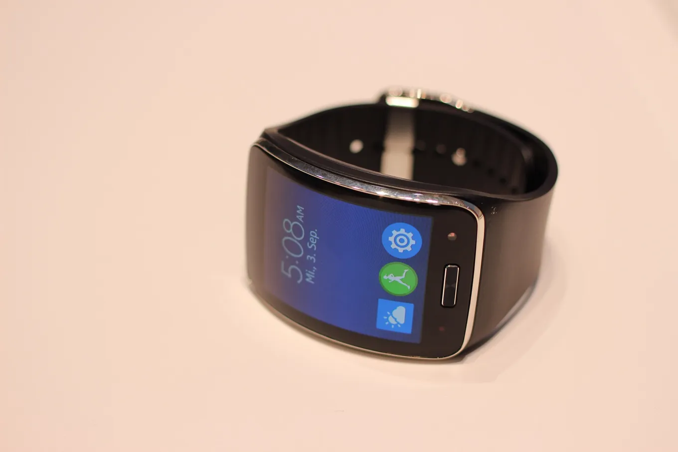 Un ceas inteligent Samsung Gear S pe o suprafață deschisă la culoare, cu ecranul curbat afișând ora și trei pictograme de aplicații.