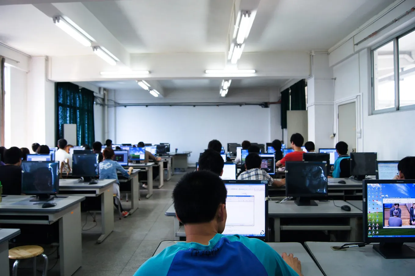Studenți care lucrează la calculatoare într-o sală de laborator universitară, văzuți din spatele sălii.