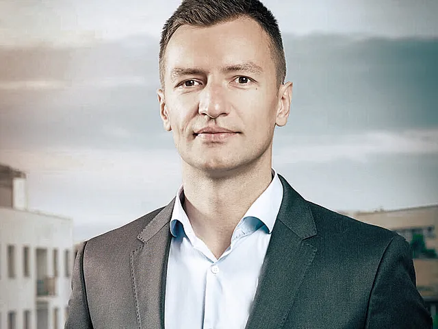 Marius Hanganu, în sacou gri închis și cămașă bleu cu gulerul descheiat, pe fundal acoperișuri și cer înnorat.