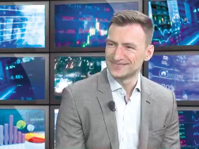 Marius Hanganu zâmbind în studioul ZF Live, în sacou gri, cu microfon la rever, pe fundalul unui perete de ecrane cu grafice bursiere.