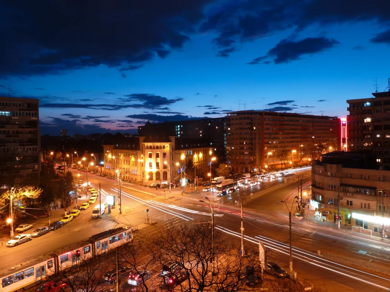 București la ora albastră, văzut de sus: o intersecție luminată, cu dâre de lumină lăsate de mașini și un tramvai, blocuri și o clădire de colț luminată, sub un cer albastru-închis cu nori.