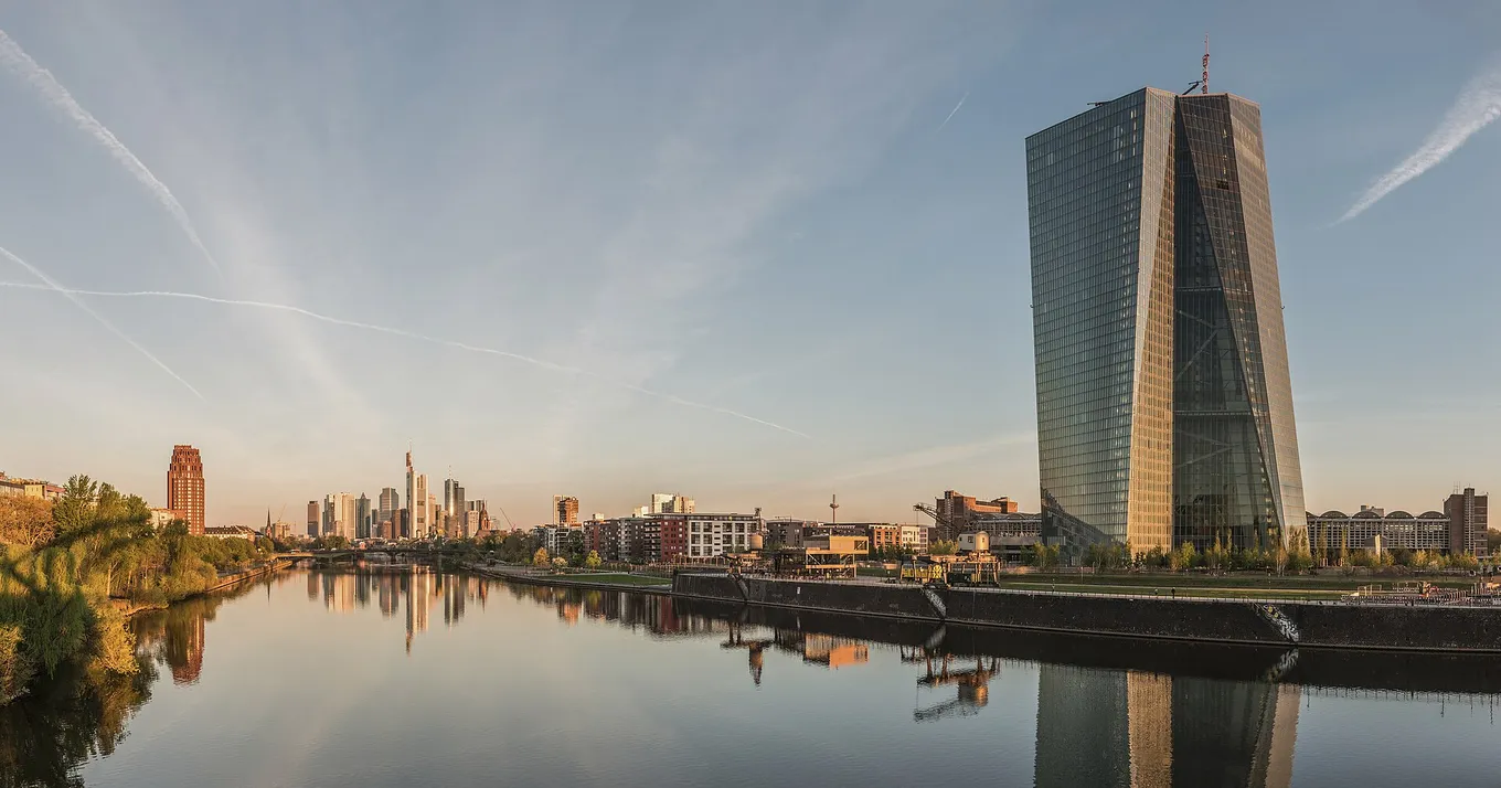 Turnul de sticlă al Băncii Centrale Europene pe malul râului Main, la Frankfurt, în zori, cu siluetele clădirilor orașului reflectate în apă, în stânga.