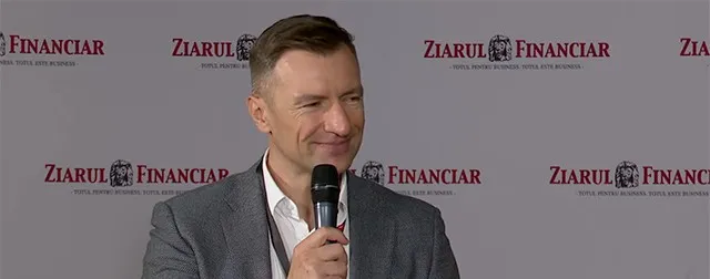Marius Hanganu zâmbind, cu microfonul în mână, în sacou gri, în fața unui panou Ziarul Financiar.